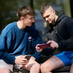 Dwóch młodych mężczyzn siedzi na ławce, jeden pokazuje drugiemu, jak przesłać zdjęcia z telefonu na telefon.