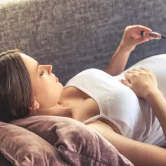 Kobieta w ciąży leży na kanapie, mierząc temperaturę. Martwi się, czy to tylko przeziębienie, czy może gorączka w ciąży.