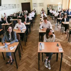 Uczniowie siedzą w ławkach podczas egzaminu, a nauczycielka stoi z tyłu.