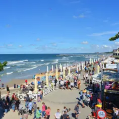 Tłum ludzi spaceruje promenadą w Kołobrzegu, obok plaży i restauracji. Zastanawiasz się, ile kosztuje klimatyczne w Kołobrzegu?