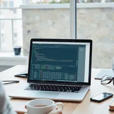 Programista pracuje przy laptopie, na ekranie widać kod. Obok leżą okulary, telefon i książka. Może właśnie używa psexec do zdalnego zarządzania.