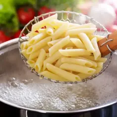 Penne makaron al dente w cedzaku nad gotującą się wodą. W tle pomidory i sałata.