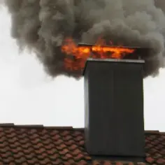 Ogień i gęsty dym wydobywają się z komina na dachu. Widać płomienie, co sugeruje, że to właśnie jest sposób, jak się gasi sadze w kominie.