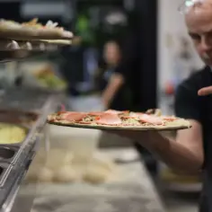 Pizzaiolo, kto to? To mistrz pizzy, który prezentuje swoje dzieło. Na pierwszym planie pizza z szynką, w tle inne gotowe.