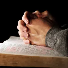 Dłonie złożone do modlitwy nad otwartą Biblią, na której widnieje tekst Składu Apostolskiego. Wierzę w Boga.