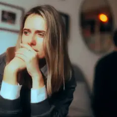 Smutna kobieta z rękami przy ustach, w tle mężczyzna odwrócony plecami. Widać obojętność w związku, cisza i dystans.