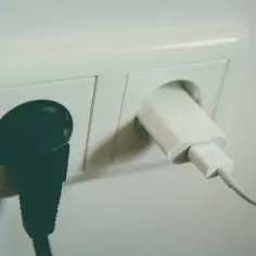 Dwa gniazdka elektryczne: jedno z czarną wtyczką, drugie z białą ładowarką. Czekają na napięcie w gniazdku.