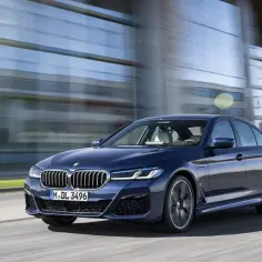 Granatowe BMW G30 540i sunie po drodze, odbijając światło od nowoczesnej fasady budynku.