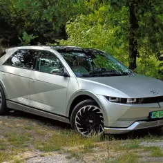 Srebrny Hyundai Ioniq 5 z czarnymi felgami stoi na kamienistej drodze, otoczony zielenią i starym budynkiem.