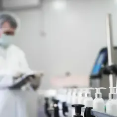 Pracownik w białej odzieży ochronnej nadzoruje linię produkcyjną z buteleczkami zawierającymi kosmetyki, gdzie kluczowym składnikiem jest fenoksyetanol.
