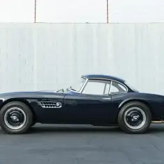 Elegancki, granatowy BMW 507 z lat 50. stoi na tle białej, ceglanej ściany.