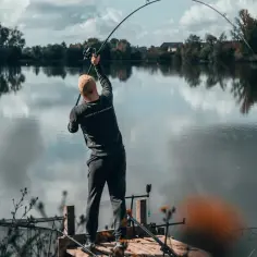 Wędkarz z zagiętą w łuk wędką, walczy z rybą. W tle spokojne jezioro i jesienne drzewa.