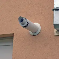 Biała rura wentylacyjna wychodząca ze ściany budynku, obok metalowego kanału.