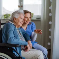 Uśmiechnięta seniorka na wózku inwalidzkim i opiekunka omawiają wniosek o świadczenie wspierające, patrząc przez okno.