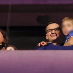 Mateusz Morawiecki z dziećmi, uśmiechnięty, trzyma na rękach jedno z dzieci. Obok kobieta patrzy w górę.