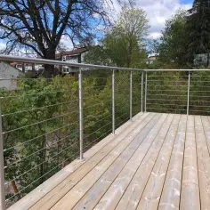 Nowe, drewniane deski tarasowe i solidne mocowanie balustrady balkonowej ze stali nierdzewnej. W tle zieleń drzew i błękitne niebo.