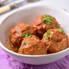 Pulpety w sosie pomidorowym bez smażenia, polane sosem i posypane natką pietruszki, podane w białej misce na fioletowej serwetce.