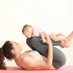 Mama i dziecko ćwiczą po porodzie na macie. Radosna mama leży na plecach, trzymając malucha na nogach.