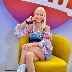 Uśmiechnięta blondynka w kwiecistej bluzce i jeansowych szortach siedzi na żółtym fotelu. Elwira Mejka, data urodzenia nieznana, pozuje do zdjęcia.