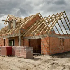 Budowa domu z czerwonej cegły z drewnianym dachem, materiały budowlane na placu.