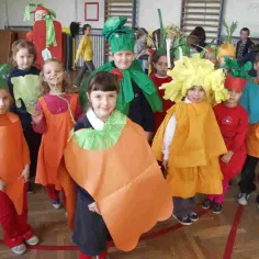 Dzieci w przedszkolu prezentują swoje przebrania za warzywa i owoce. Wśród nich marchewki, dynia i inne kolorowe stroje.