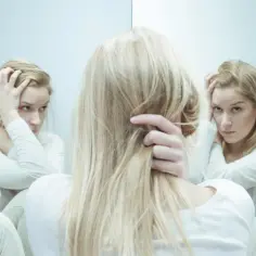 Kobieta z blond włosami w białej bluzie, z rękami na głowie, otoczona odbiciami w lustrze, symbolizującymi walkę z nerwicą lękową, być może szukająca ulgi w kwetaplex.