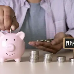 Oszczędzanie na emeryturę pomostową: ręka wrzuca monetę do świnki skarbonki, obok stosy monet i tabliczka z napisem "PENSION".