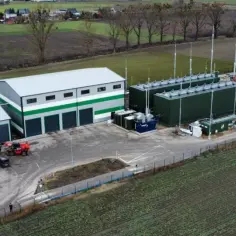Nowoczesna biometanownia z zielonymi zbiornikami i kopułą, otoczona polami i budynkami gospodarczymi.