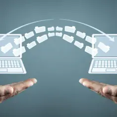 Dwie dłonie podtrzymują symboliczny transfer danych między laptopami. Proces ten, przypominający dekretację, ilustruje wymianę informacji.
