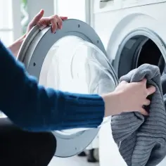Ręce wkładają szary sweter do pralki, by rozwiązać problem, jak pozbyć się kłaczków i sierści psa z ubrań w trakcie cyklu prania.