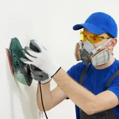 Pracownik w masce i okularach ochronnych wykonuje szlifowanie gładzi na białej ścianie za pomocą szlifierki.