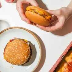 Ręce trzymają burgera z serem, obok talerz z drugim burgerem i kawałek pizzy. Idealne pytanie: jakie mięso do burgera wybrać?