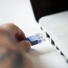 Dłoń z tatuażami podłącza niebieski kabel sieciowy do portu Ethernet w białym laptopie.
