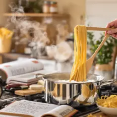 Dłoń trzyma drewniane szczypce z makaronem spaghetti nad garnkiem z gotującą się wodą.