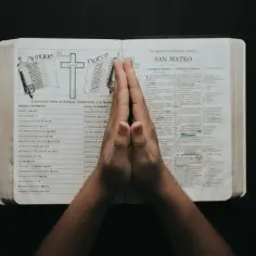 Dłonie złożone do modlitwy nad otwartą Biblią, na stronach której widnieje porównanie Starego i Nowego Testamentu, a także fragment Ewangelii wg św. Mateusza. W tle widać symbol krzyża i zwoju, nawiązujące do treści. To chwila refleksji, być może nad p...