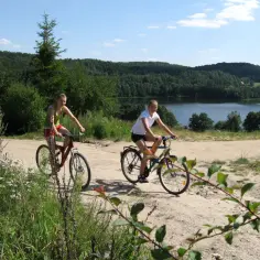 Dwie dziewczyny na rowerach przemierzają kaszubską marszrutę, podziwiając jezioro i las.