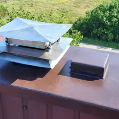 Metalowy daszek na komin z blachy na brązowym dachu, obok mniejszej brązowej osłony.