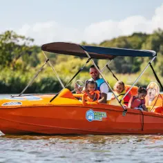 Rodzina na pomarańczowej łódce z napisem "Water Fun" płynie po jeziorze. Ojciec z dziećmi cieszą się słońcem, idealny dzień na wodne atrakcje w Mielnie.