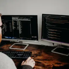 Mężczyzna z brodą i okularami pracuje przy dwóch monitorach z kodem. Czy szukanie pracy w IT bez doświadczenia, czy sam bootcamp wystarczy?
