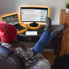 Kobieta w czerwonej czapce i szarej kurtce siedzi przy biurku, z nogami na blacie, przed laptopem i monitorem.