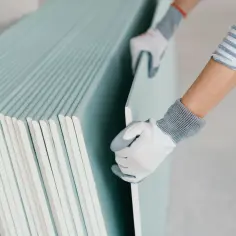 Ręce w rękawiczkach chwytają płytę karton gips. Widoczna grubość materiału budowlanego.