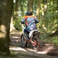 Motocyklista w kasku i pomarańczowo-szarej bluzie jedzie po leśnym torze. Od ilu lat można jeździć crossem? Odpowiedź zależy od przepisów i umiejętności.