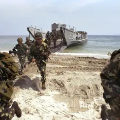 Żołnierze w pełnym rynsztunku, z karabinami w rękach, biegną po plaży. Za nimi widać okręt desantowy, z którego właśnie wyszli. To dynamiczny desant wojsk.
