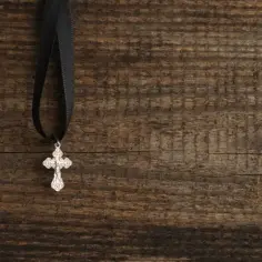 Srebrny naszyjnik z krzyżem na czarnej wstążce, zawieszony na drewnianym tle.