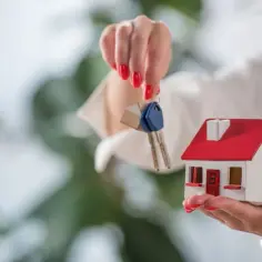 Kobieta trzyma w dłoniach miniaturowy domek i klucze. To symboliczne przedstawienie, jak mieszkanie może stanowić wkład własny przy zakupie nieruchomości.