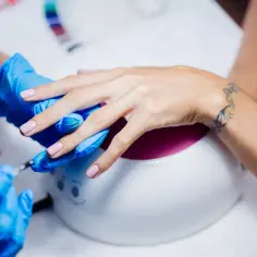 Manicure w trakcie: dłonie w niebieskich rękawiczkach, lampa UV, paznokcie żelowe. Kolejnosc robienia paznokci zelowych.