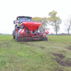 Rolnik aktywny zawodowo pracuje na polu, używając czerwonej siewnicy połączonej z traktorem.