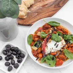 Pyszna burrata z pieczonymi pomidorkami koktajlowymi, czosnkiem i bazylią, skropiona octem balsamicznym. Obok czarne oliwki i kromki chleba.