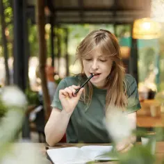 Młoda kobieta zamyślona nad notatkami, gryzie długopis. Może pisze esej "for and against", szukając argumentów.