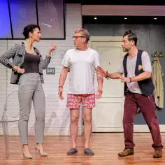 Trzech aktorów na scenie. Kobieta w eleganckim żakiecie i spodniach gestykuluje, mężczyzna w białej koszulce i szortach z kotami patrzy na nią z niedowierzaniem, a trzeci mężczyzna w kamizelce i bordowych spodniach próbuje ich pogodzić. Lekko nie będzi...